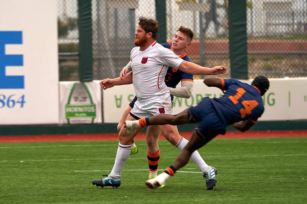 Rugby players in blue trying to stop a player wearing white.