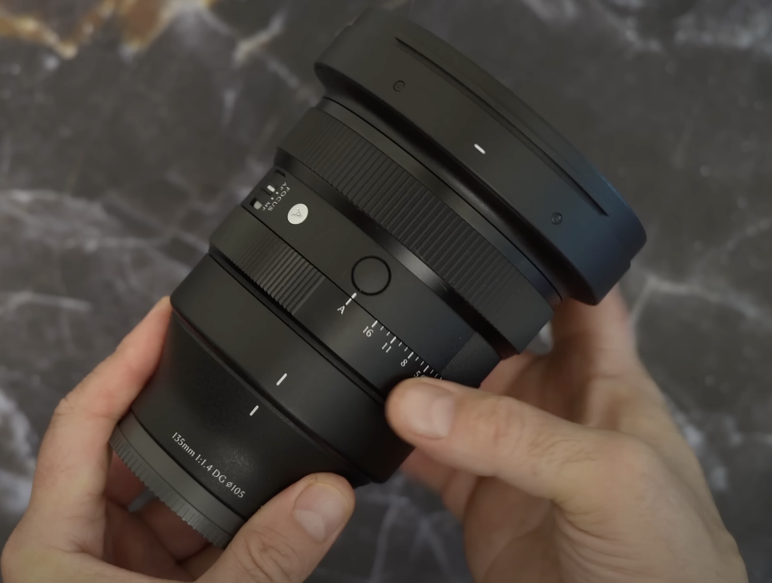 The Sigma 135mm f/1.8 Art lens being held over a marble surface. Aperture ring is visible. 