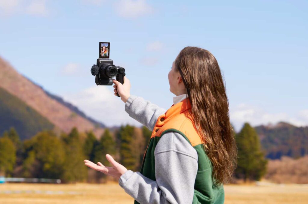 A woman with long hair holding the Sony ZV-E10 in a clearing near a mountain
